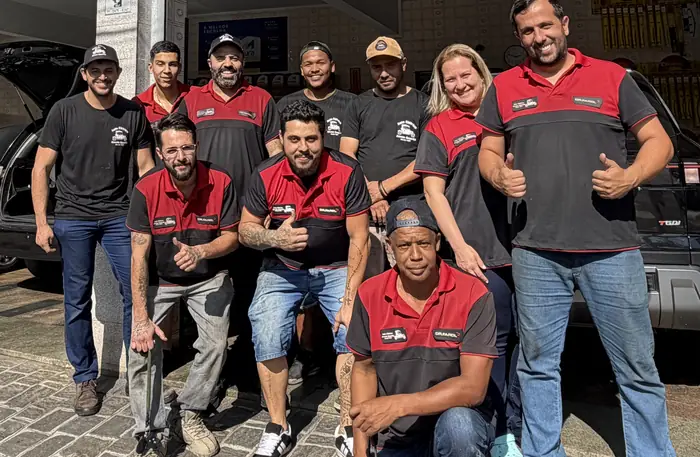 Equipe do Auto Elétrico Jovem Guarda, dez pessoas com o uniforme vermelho e preto da oficina, em frente à fachada com o letreiro